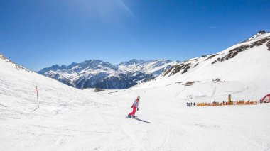 A snowboarder going down the slope in Heiligenblut, Austria. Perfectly groomed slopes. High mountains surrounding the girl, wearing pink trousers and colorful jacket. Girl wears helm for protection