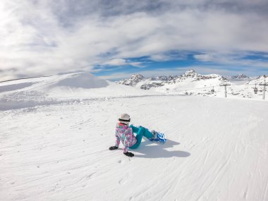 A snowboarder sitting on the slope in Moelltaler Gletscher, Austria. Perfectly groomed slopes. High mountains surrounding the girl wearing colourful snowboard outfit. Girl wears helm for the protection