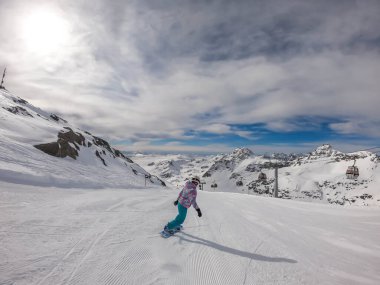 A snowboarder going down the slope in Moelltaler Gletscher, Austria. Perfectly groomed slopes. High mountains surrounding the girl wearing colourful snowboard outfit. Girl wears helm for the protection
