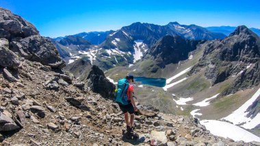 A young woman with a big backpack hikes down towards a clear, navy blue lake hiding between tall mountain peaks. Some of the slopes are covered with snow. In the back is another mountain range