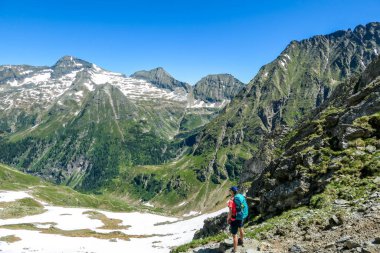 A young woman with a big backpack hikes down on a steep pathway between tall mountain peaks. Some of the slopes are covered with snow. In the back is another mountain range. Spring in alpine valleys.