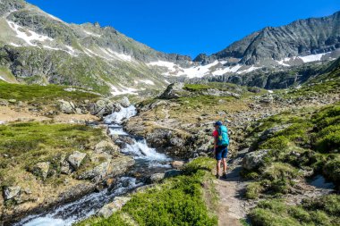 A young woman with a big backpack hikes up on a steep pathway, along a mountain stream. the water cascades on the rocks, rushing into the valley. Girl is enjoying the spring in the mountains