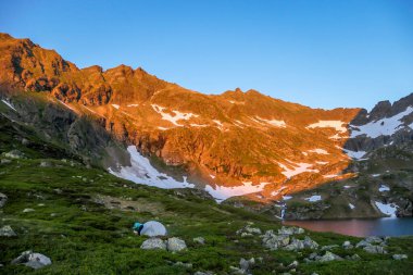 A woman camping in a wilderness. She puts up a small tent, placed in a vast valley, with beautiful sunset in the mountain peaks. High mountains around. Spring in alpine valleys. Calmness and happiness