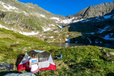 A woman camping in a wilderness. She sits next to a small tent, placed on a top of a mountain peak, waiting for the sunset. High mountains around. Spring in alpine valleys. Calmnes and happiness.