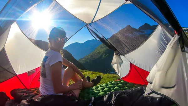 A woman camping in a wilderness. She sits in a small tent, placed on a top of a mountain peak, waiting for the sunset. High mountains around. Spring in alpine valleys. Calmnes and happiness.