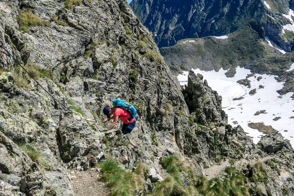 A young woman with a big backpack hikes down a steep slope using a metal rope. Girl is focused and concentrated. Dangerous hike. Steep via ferrata.There is a lot of snow in the valley below.