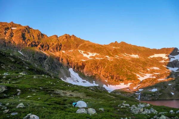 A woman camping in a wilderness. She puts up a small tent, placed in a vast valley, with beautiful sunset in the mountain peaks. High mountains around. Spring in alpine valleys. Calmness and happiness