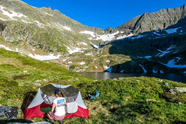 A woman camping in a wilderness. She sits next to a small tent, placed on a top of a mountain peak, waiting for the sunset. High mountains around. Spring in alpine valleys. Calmnes and happiness.