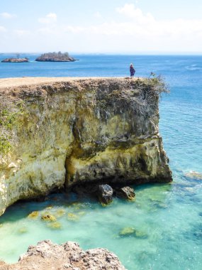A woman in maxi dress walking at the edge of cliffs of Pink Beach in Lombook, Indonesia. The girl is wearing a straw hat. The sea water has clear turquoise colour. Happiness and adventurous travelling