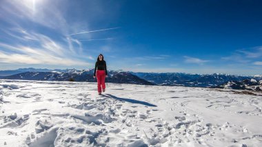 A girl with a snowboarding outfit standing on a powder snow, off the trail in Bad Kleinkirchheim, Austria. There are few traits on the snow. Girl is smiling, enjoying beautiful weather and view.