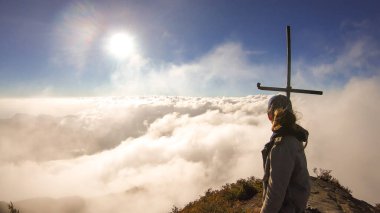 A young woman standing on the top of volcano Inerie in Bajawa, Flores, Indonesia, next to a cross. She is surrounded by thick clouds. Sun tries to get through the thick layer of overcast. Freedom