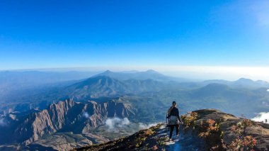 A young woman walking down the volcano Inierie in Bajawa, Flores, Indonesia. She supports herself on a wooden stick while enjoying the beautiful view on volcanic island. Lots of fluffy clouds around