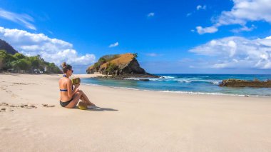 A girl in bikini drinking fresh coconut juice, while sitting on the idyllic Koka Beach and watching the calm sea. Hidden gem of Flores, Indonesia. Island life. Happiness and simplicity. Healthy diet.