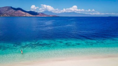 A drone shot of a girl playing on the beach on a small island near Maumere, Indonesia. Happy and careless moments. The coast changes colors from white to turquoise and navy blue. There are few islands