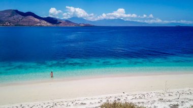 A drone shot of a girl playing on the beach on a small island near Maumere, Indonesia. Happy and careless moments. The coast changes colors from white to turquoise and navy blue. There are few islands