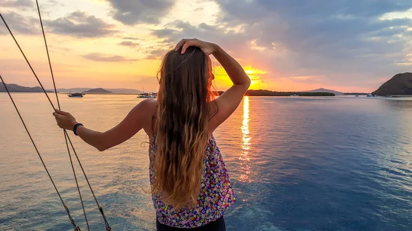 A girl with long hair on a small boat enjoying beautiful sunset in Komodo National Park, Flores, Indonesia. Sun sets over the horizon line. Girl is holding her hair in her hand, trying to tame it
