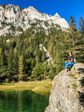 Girl sitting on a big boulder at the shore of Green Lake in an Alpine valley in Austria and enjoying the peaceful mountain view. Spring in the valley. Dense forest in the back. Playtime and happiness.