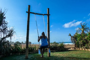A girl swinging on a bamboo swing with the view on Nyang Nyang Beach, Bali, Indonesia. The swing has very simple construction. Hidden getaway. Collecting happy moments and having fun.