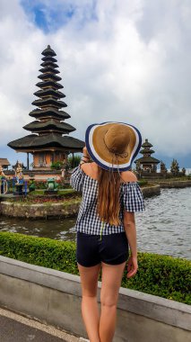 A woman in shorts and straw hat standing in front of main building of Ulun Danu Temple, Bali, Indonesia. The girl is admiring the beauty of Hindu water temple. Sacred place. Discovering new cultures.