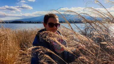 A girl walking between golden grass at the shore of Kawaguchiko Lake and the view on Mt Fuji in the back. The mountain hides behind the clouds. She is enjoying her time, exploring the unbeaten paths