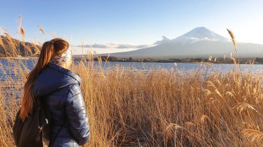A woman standing in between golden grass at the shore of Kawaguchiko Lake, Japan with the view on Mt Fuji. The mountain surrounded by clouds. Soft reflections in the calm surface of the lake. Serenity