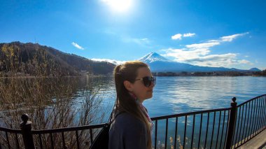 Woman walking across the bridge next to Kawaguchiko Lake, Japan with the view on Mt Fuji. The mountain is surrounded by clouds. Girl is enjoying the walk and good weather. Serenity and calmness