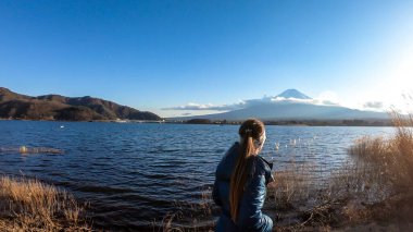 A woman walking in between golden grass at the shore of Kawaguchiko Lake, Japan with the view on Mt Fuji. The girl is enjoying the view on the volcano. The mountain surrounded by clouds. Serenity
