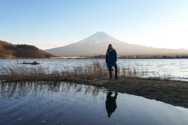 Kawaguchiko Gölü 'nün kenarında yürüyen ve Japonya' daki Fuji Dağı 'nı izleyen bir kadın. Sudaki kadının yansıması gibi. Volkanın tepesi karla kaplı. Yeni yerler keşfetmek. Huzur. Yumuşak günbatımı renkleri