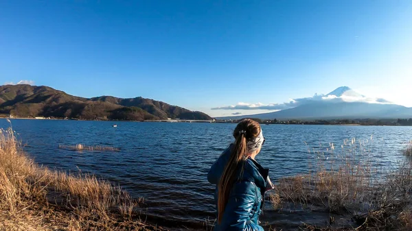 A woman walking in between golden grass at the shore of Kawaguchiko Lake, Japan with the view on Mt Fuji. The girl is enjoying the view on the volcano. The mountain surrounded by clouds. Serenity