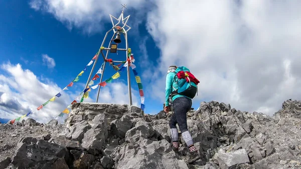 A woman reaching the top of Monte Coglians, Hohe Warte on the Austrian-Italian Alpine border. Metal construction with a bell, wrapped with prayer flags. She is not giving up, visible goal. Overcast