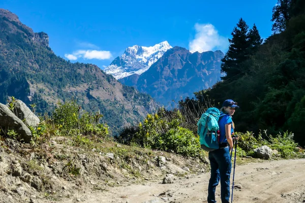 A girl with a big blue backpack hikes along a pathway on Annapurna Circuit Trek in Himalayas, Nepal. There are mountain chains around her. The slopes are overgrown with green plants. Discovering