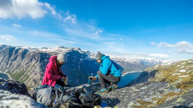 A couple having breakfast in the tall mountains with a view on a fjord-like lake. Girl is cutting the bread and the boy boils the water. Clear and sunny weather. Camping in the wilderness.