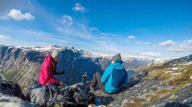 A couple having breakfast in the tall mountains with a view on a fjord-like lake. They are both relaxed and chilled. Clear and sunny weather. Camping in the wilderness in beautiful nature.
