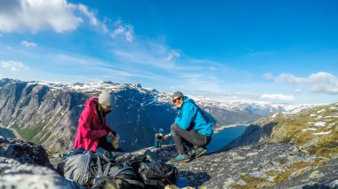 A couple having breakfast in the tall mountains with a view on a fjord-like lake. Girl is cutting the bread and the boy boils the water. Clear and sunny weather. Camping in the wilderness.
