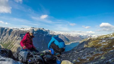 A couple having breakfast in the tall mountains with a view on a fjord-like lake. Girl is holding a pink cup and the boy boils the water. Clear and sunny weather. Camping in the wilderness.