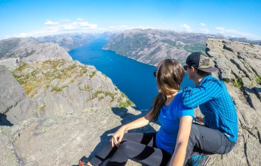 A couple sitting at the edge of a steep cliff of Preikestolen, with a view on Lysefjorden. Fjord goes far inland. The couple enjoys the view, feeling free and happy. Great accomplishment