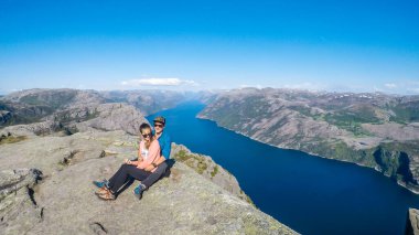 A couple sitting at the edge of a steep cliff of Preikestolen, with a view on Lysefjorden. Fjord goes far inland. The couple enjoys the view, feeling free and happy. Great accomplishment