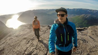 Couple walking close to the edge of a steep cliff of Preikestolen, with a view on Lysefjorden. Sun slowly goes down, reflecting itself in the water. The couple enjoys the view, feeling free and happy