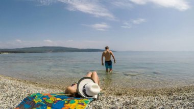 A couple on the beach. The girl is lying on a mat and suntanning, whilst the man is going into the sea. Couple travels. Stony beach and turquoise color of the water. Clear and sunny day.