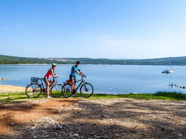 A couple in sporty outfits stand on their bikes by the seashore and admire the view behind them. Coast is partially sandy, stony and grassy. The water in the bay is calm, boat anchored to the shore