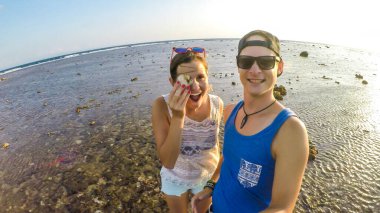 Senggigi plajında oynayan çift, Lombok, Endonezya. Kızın elinde gözünü kapatan bir deniz kabuğu var. Adam selfie çekiyor. Sığ bir deniz suyunda duruyorlar. Seyahat ederken beraberlik ve sevgi