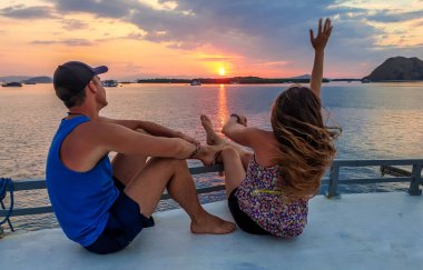 Couple sitting on a small boat enjoying beautiful sunset in Komodo National Park, Flores, Indonesia. Sun sets over the horizon line. They are having fun, sailing across the calm sea. Love and romance