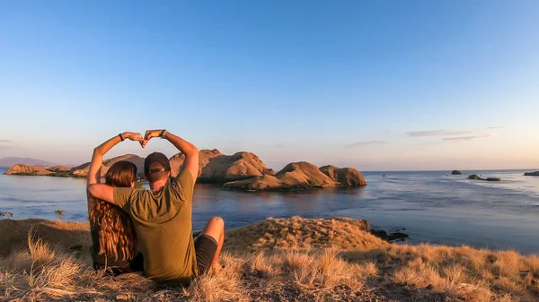 一对夫妇坐在一个小岛上 在印度尼西亚弗洛雷斯科莫多国家公园的上空享受晨阳 岛上和海上的黄金时刻 他们用自己的双手形成了一颗心 — 图库照片