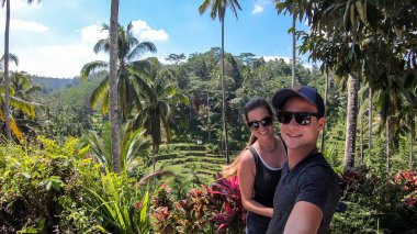 A couple taking a selfie with rice terraces shining in bright green colors in Ubud, Bali, Indonesia. Endless paddies of rice, spreading on great distances. There are palm trees between the terraces.