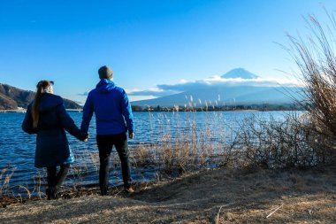 A couple holding hands at the side of Kawaguchiko Lake, Japan with the view on Mt Fuji. The mountain surrounded by clouds, top of it covered with snow. Exploring new places. Love and affection