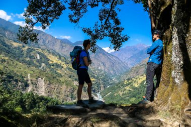 A couple in hiking outfits standing under a thick tree trunk and enjoying the view on lush green Himalayan valley along Annapurna Circuit in Nepal. They are happy and full of energy. Cheerful moments