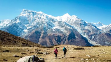A couple walking on the Annapurna Circuit Trek, Himalayas, Nepal. Snow caped Annapurna chain in the back. Clear weather, dry grass, snowy peaks. High altitude, massive mountains. Freedom and adventure