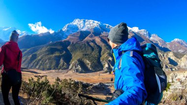 A couple trekking along Annapurna Circuit Trek, Himalayas, Nepal. Snow caped Annapurna chain in the back. They are surrounded with dried grass. Harsh landscape. Adventure and exploration.