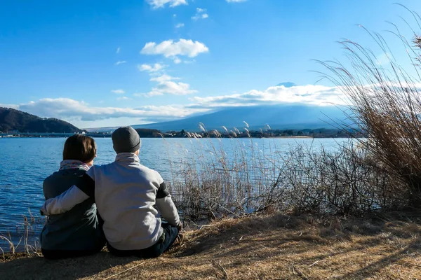 A couple sitting on the ground and watching Mt Fuji from Kawaguchiko Lake, Japan. The mountain surrounded by clouds, top of it covered with snow. Exploring new places. Love and affection.