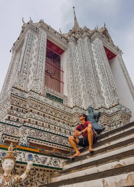 Wat Arun in Bangkok - Temple of Dawn, Thailand.  A young man sitting on the stairs of the temple. Calm and peaceful feeling. Detailed wall of the temple, mosaic patterns in white, red and green. 